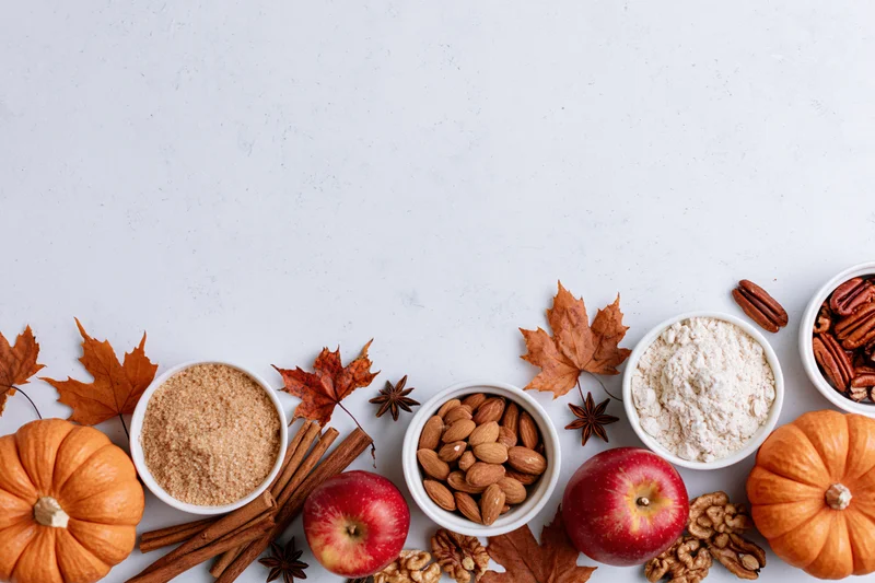 Autumn ingredients arranged in a flat lay on a white surface with pumpkins, apples, nuts, spices and maple leaves