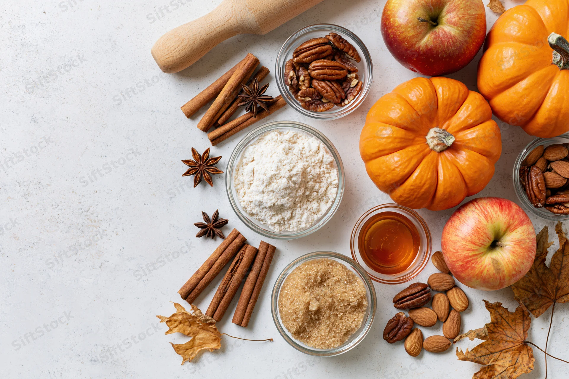 Autumn baking ingredients arranged on a white countertop with pumpkins, apples, cinnamon, nuts and spices