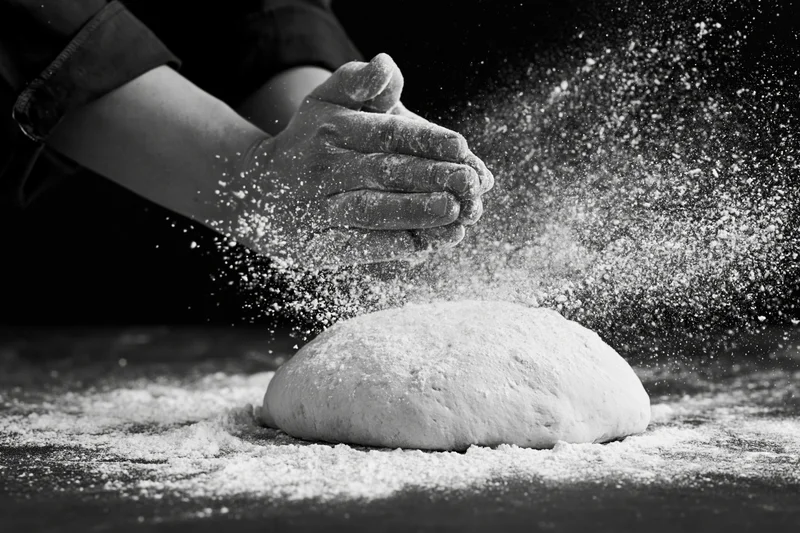 Hands clapping flour over a ball of dough on a dark surface