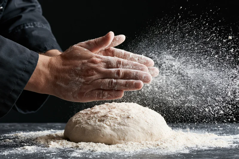Hands sprinkling flour over dough on dark surface