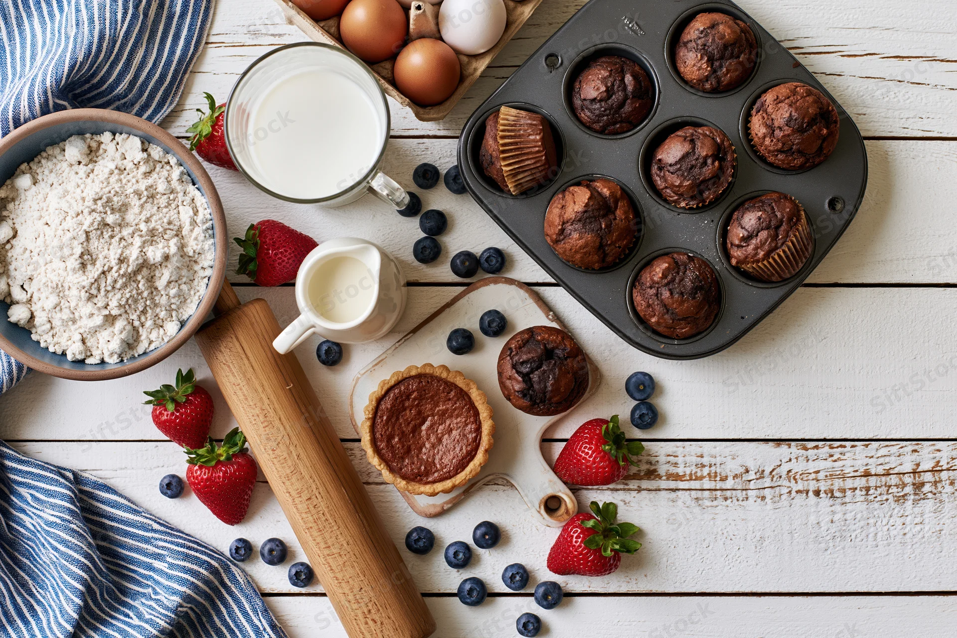 Chocolate muffins and baking ingredients arranged on a white wooden table with strawberries and blueberries
