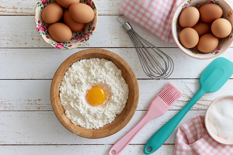 Flour in a wooden bowl with a cracked egg on top surrounded by baking tools and ingredients on a white wooden table
