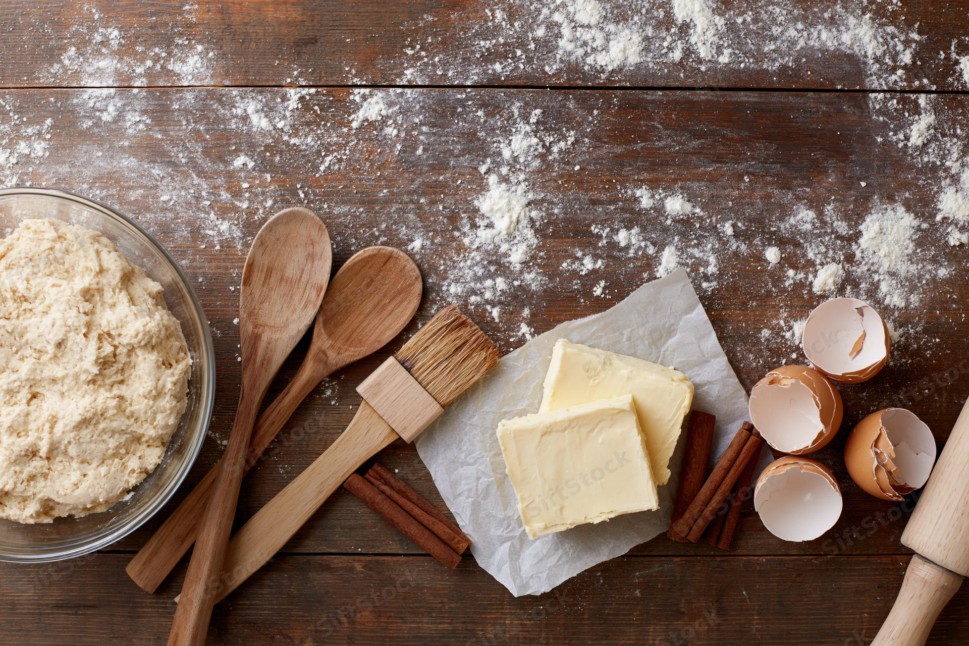Dough in a bowl with butter, eggshells, cinnamon and wooden utensils on a floured wooden table