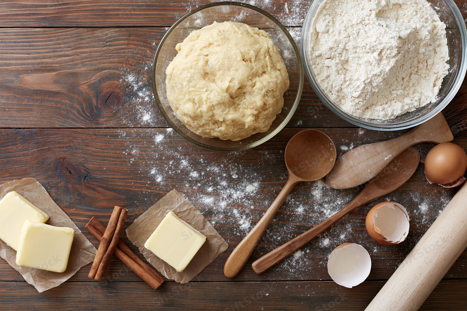 Dough and baking ingredients arranged on a wooden countertop with utensils and scattered flour