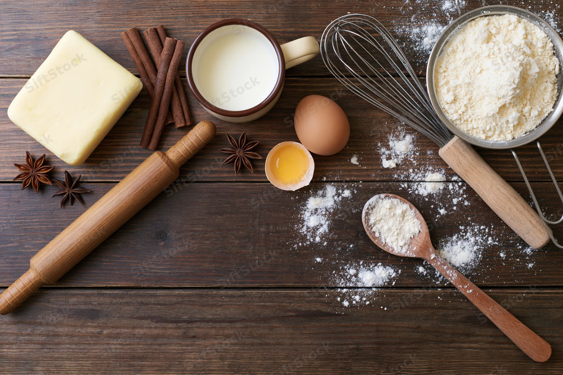 Baking ingredients arranged on a wooden table with rolling pin, whisk, and spoon
