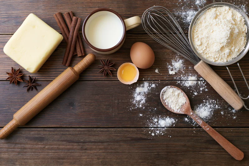 Baking ingredients arranged on a wooden table with rolling pin, whisk, and spoon