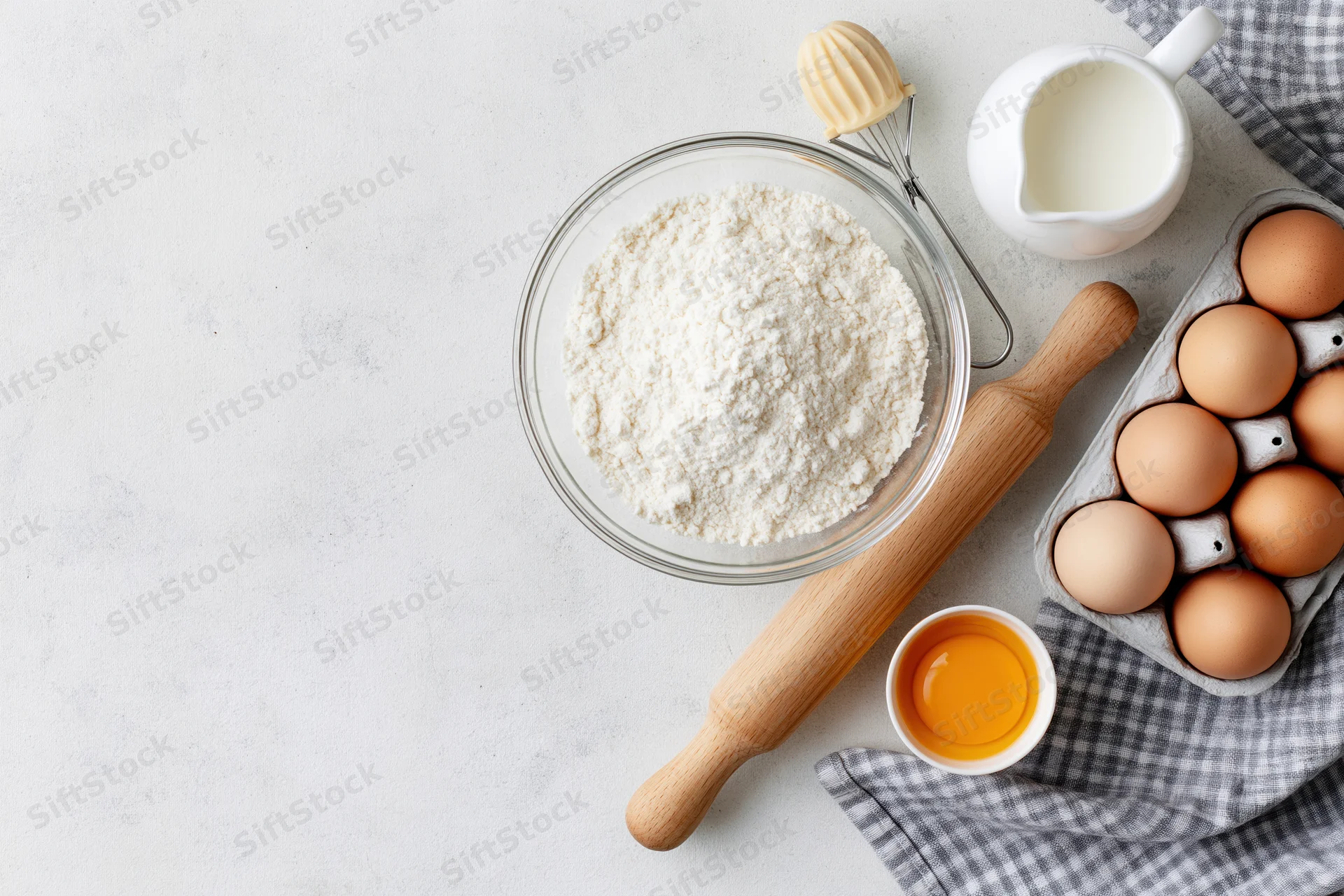 Baking ingredients arranged on a white countertop with a rolling pin and whisk, including flour, eggs, milk, and a checkered cloth