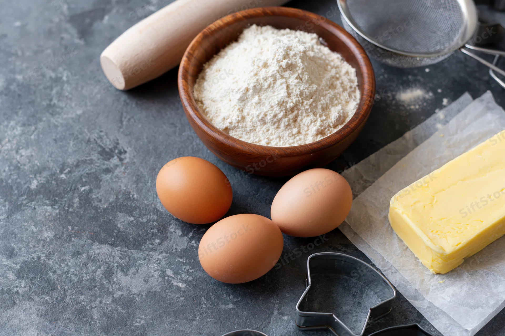 Baking ingredients including flour in a wooden bowl, eggs, butter on parchment, and cookie cutters arranged on a gray countertop