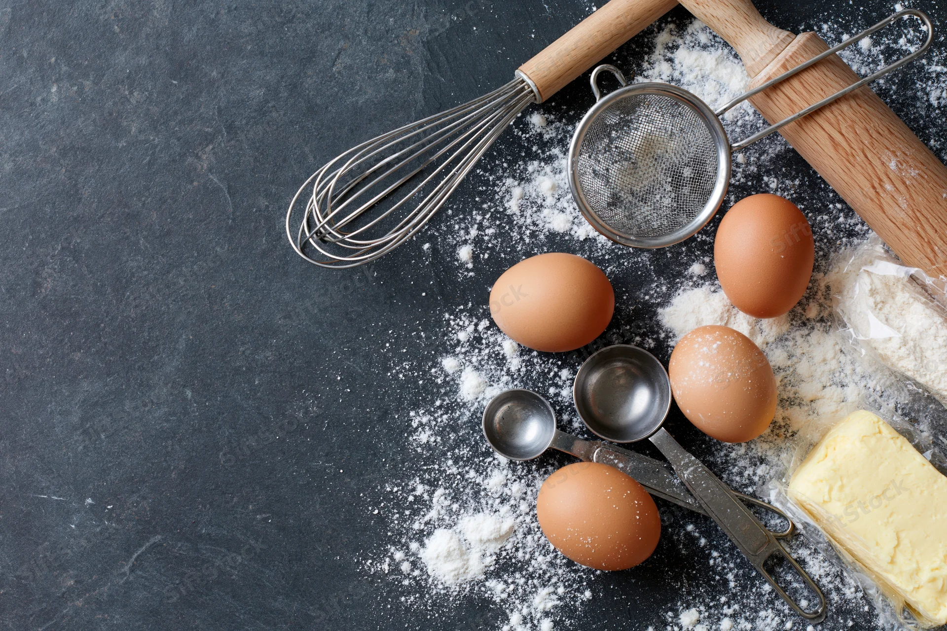 Baking ingredients including eggs, flour, butter, whisk and rolling pin arranged on dark countertop with copyspace
