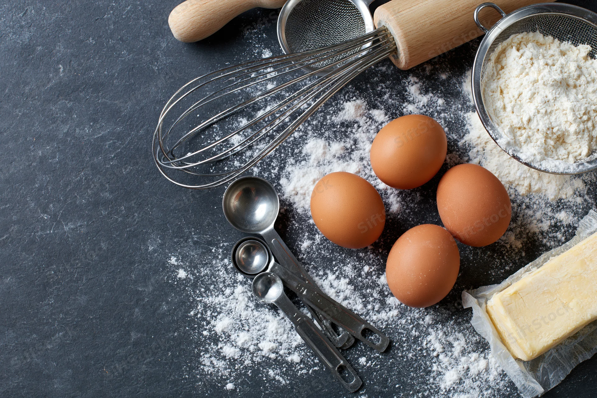 Baking ingredients including flour, eggs, butter, whisk and measuring spoons arranged on dark countertop with copyspace
