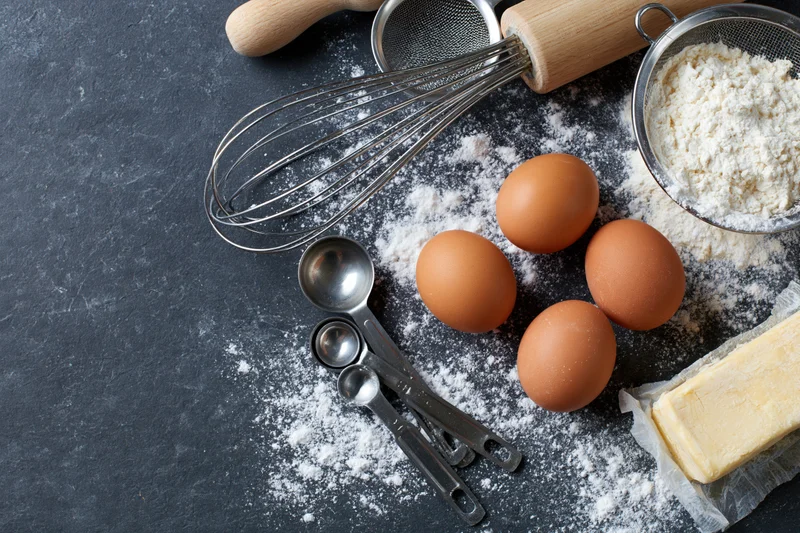 Baking ingredients including flour, eggs, butter, whisk and measuring spoons arranged on dark countertop with copyspace