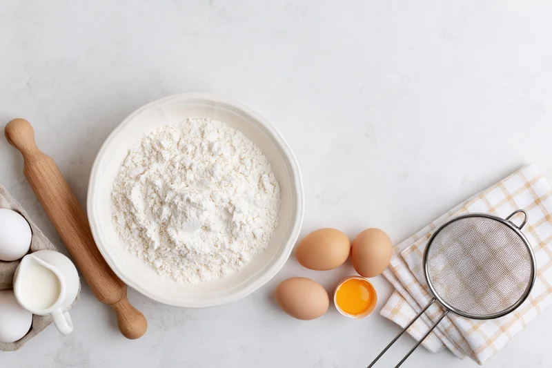 Baking ingredients arranged on a white countertop with flour in a bowl, eggs, milk, rolling pin and sieve nearby