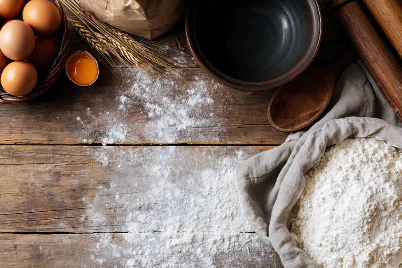 Flour scattered on a wooden table with eggs, a bowl, rolling pin and sack of flour for baking