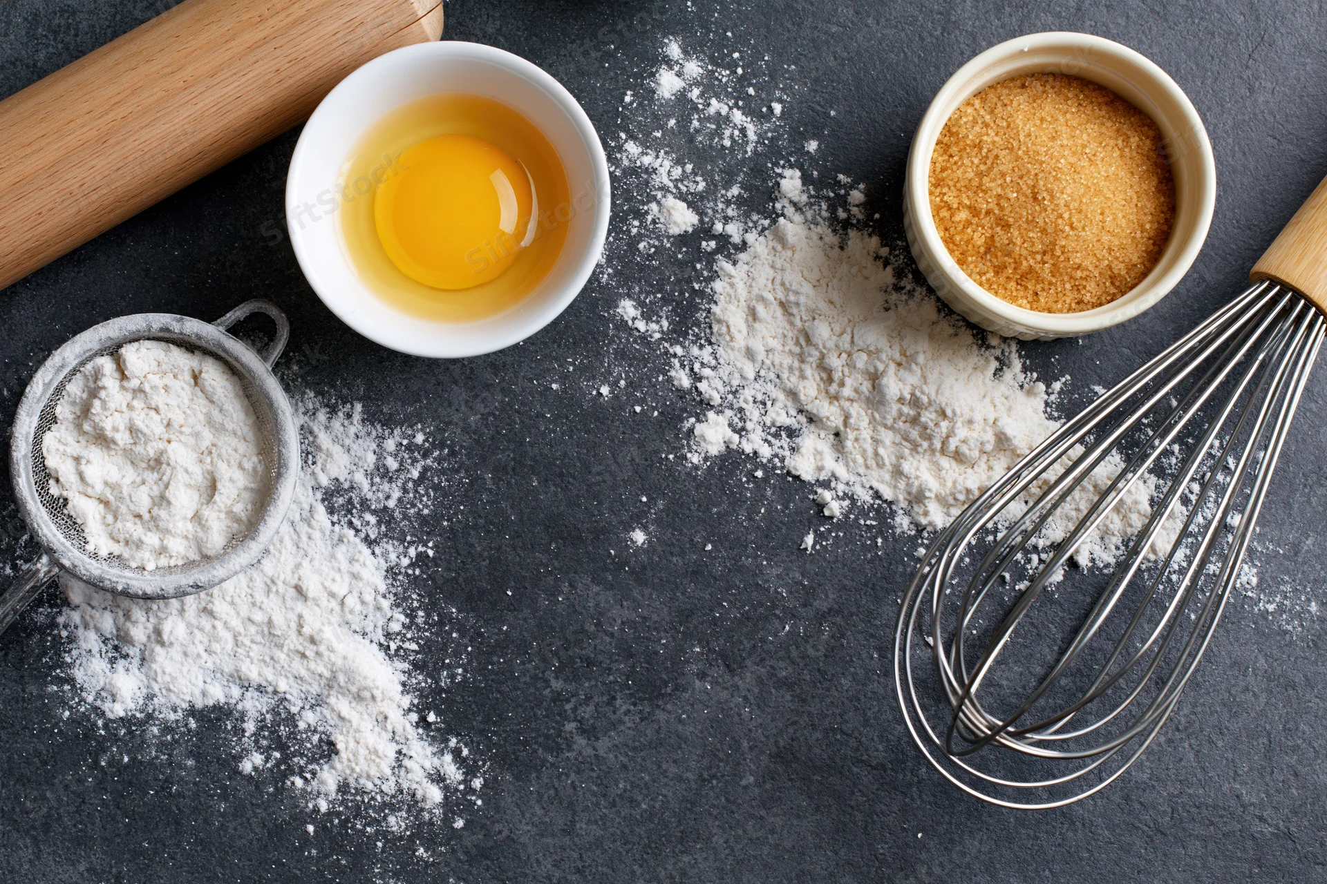 Baking ingredients arranged on dark countertop with flour, egg, sugar, whisk and rolling pin