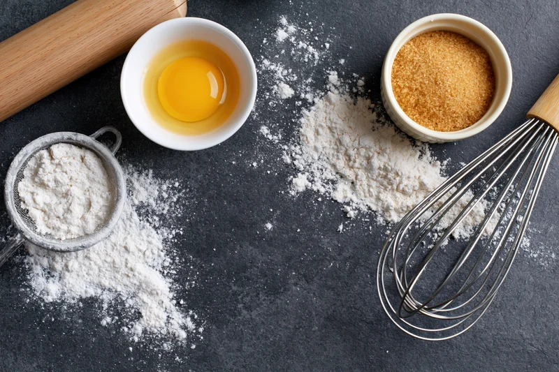 Baking ingredients arranged on dark countertop with flour, egg, sugar, whisk and rolling pin