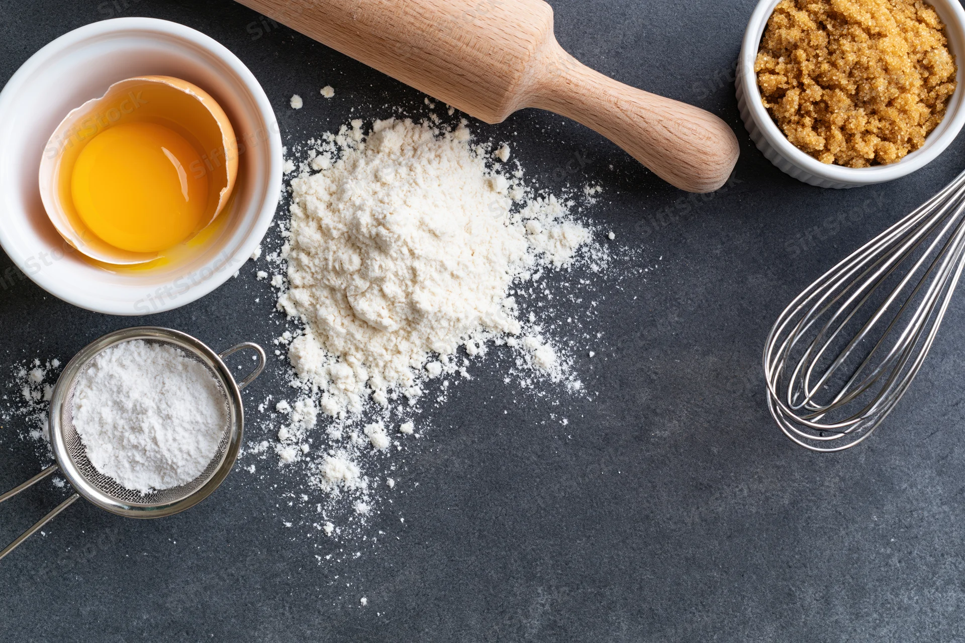 Baking ingredients arranged on dark countertop with rolling pin, whisk, and sifter
