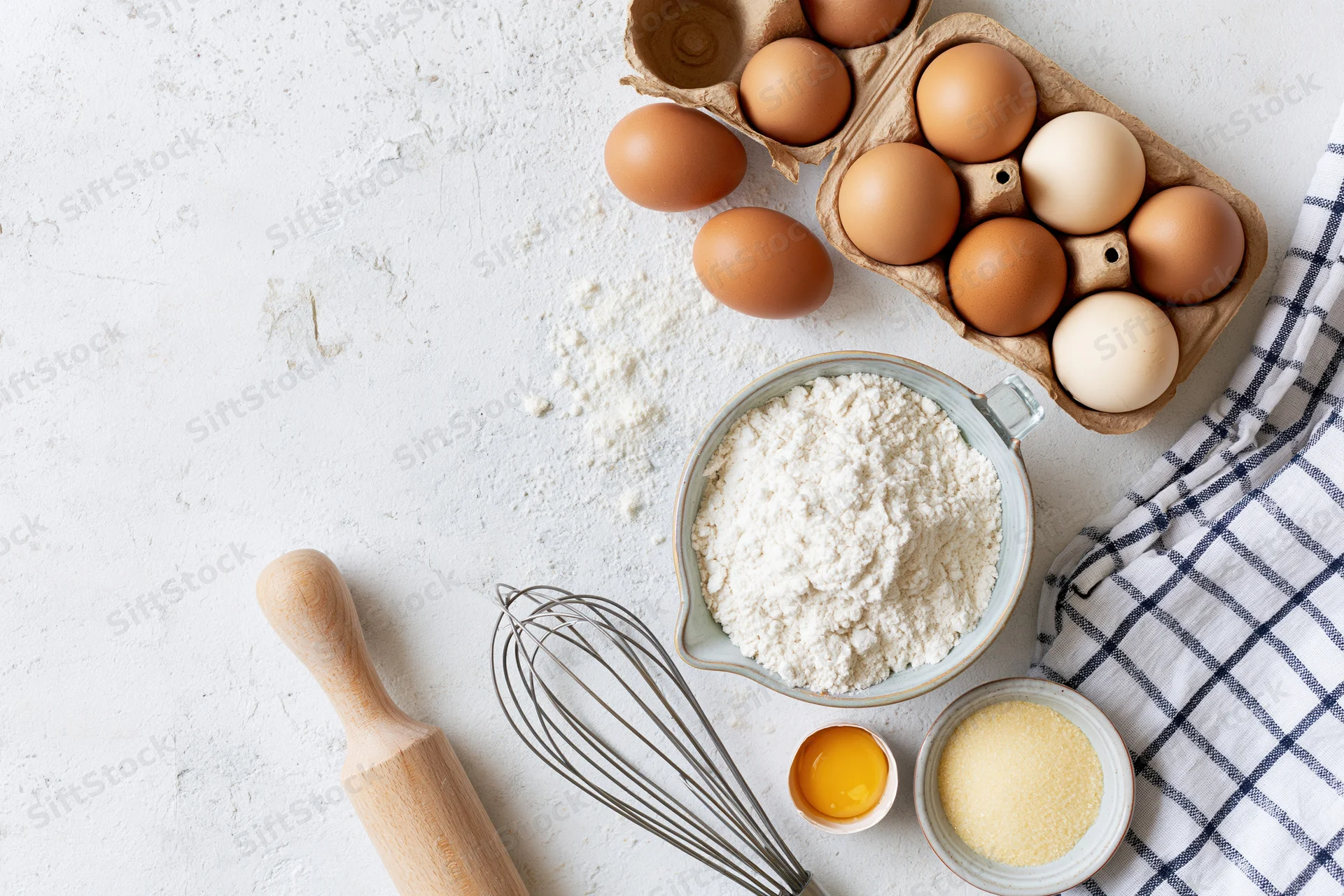 Baking ingredients arranged on a white countertop with a whisk, rolling pin and checkered cloth
