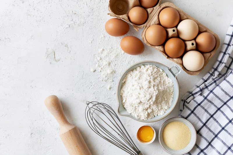 Baking ingredients arranged on a white countertop with a whisk, rolling pin and checkered cloth