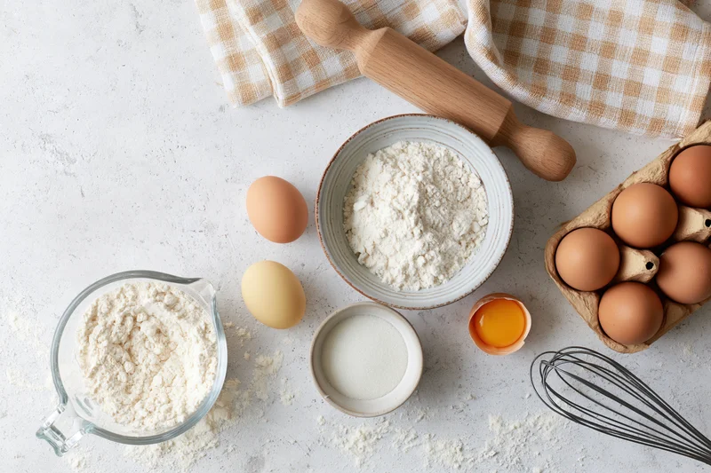 Baking ingredients arranged on a white countertop with rolling pin and whisk