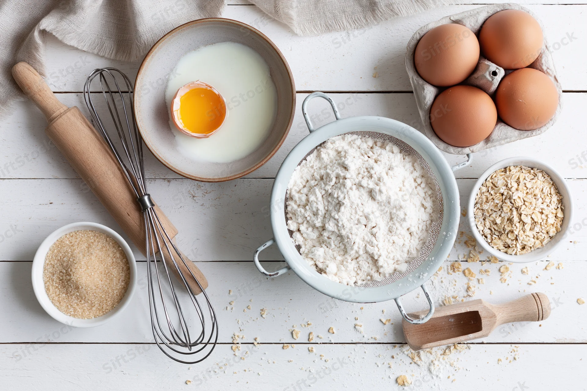 Baking ingredients arranged on a white wooden table with flour in a sifter, eggs in a carton, and utensils nearby