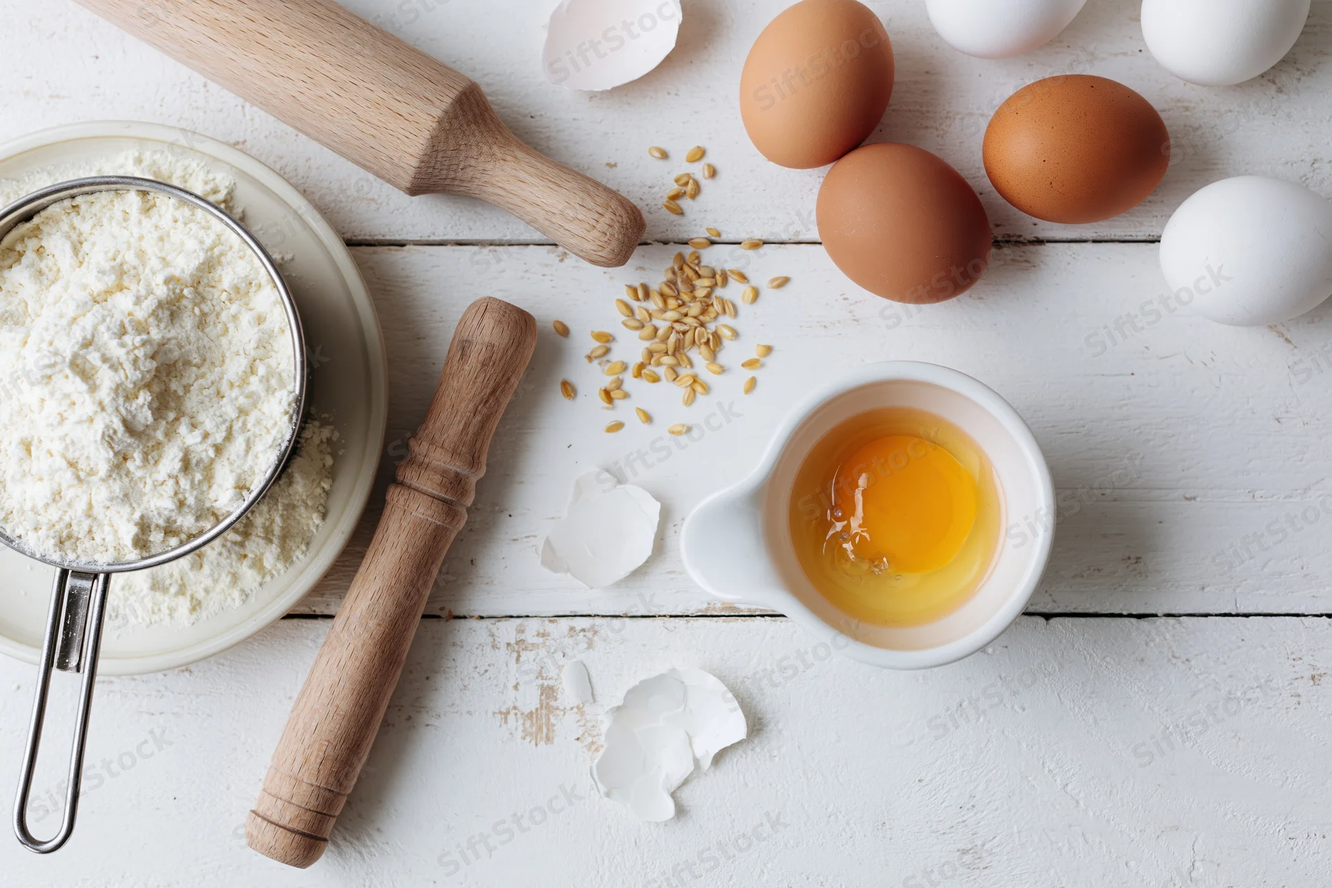 Baking ingredients including flour, eggs, and wooden tools arranged on a white wooden surface