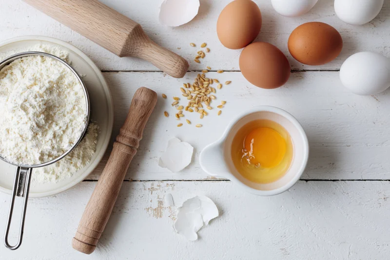 Baking ingredients including flour, eggs, and wooden tools arranged on a white wooden surface