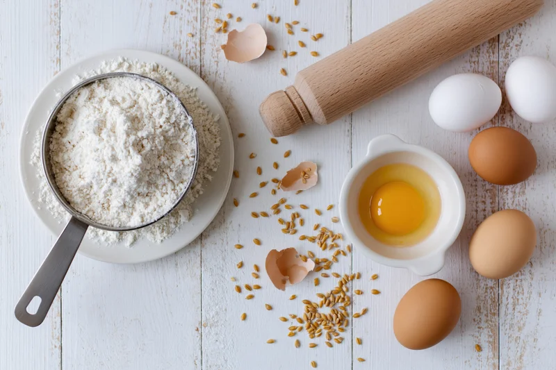 Baking ingredients including flour, eggs, and a rolling pin arranged on a white wooden table