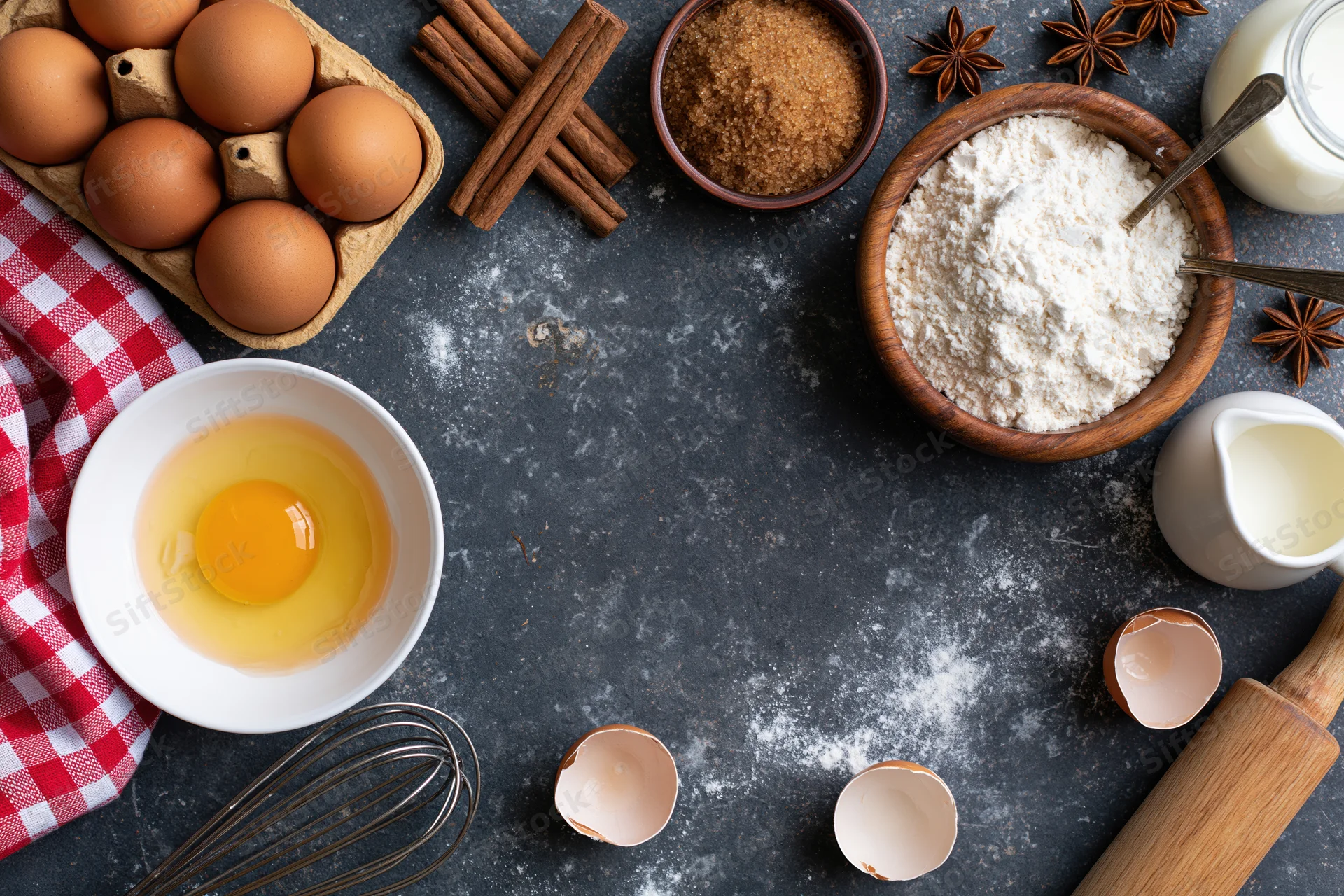 Baking ingredients arranged on dark countertop with cracked eggs and utensils