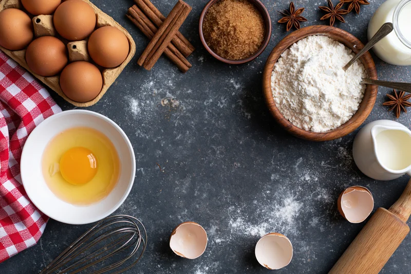 Baking ingredients arranged on dark countertop with cracked eggs and utensils