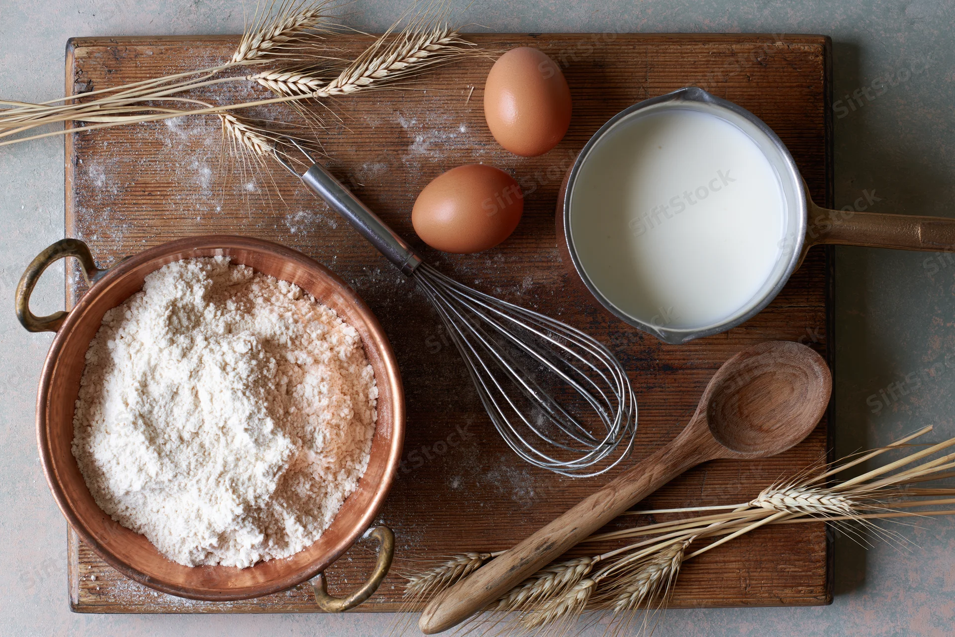Baking ingredients arranged on a wooden board with flour, eggs, milk, whisk and wheat stalks