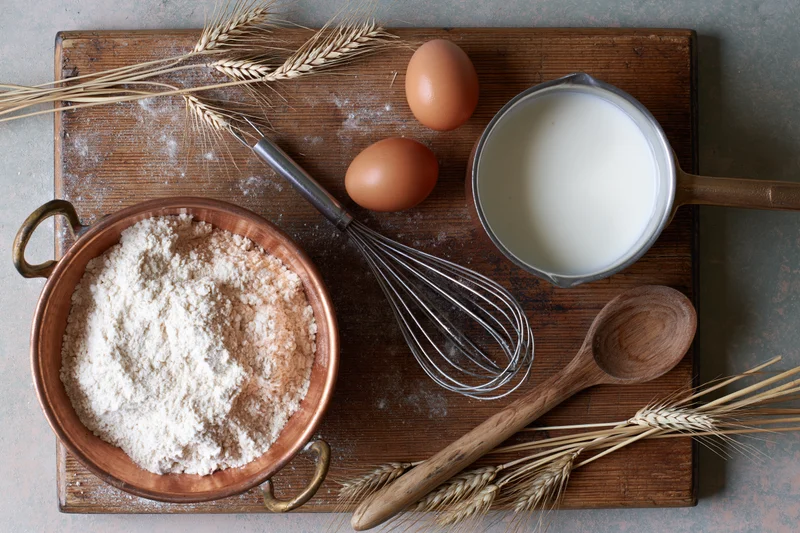 Baking ingredients arranged on a wooden board with flour, eggs, milk, whisk and wheat stalks