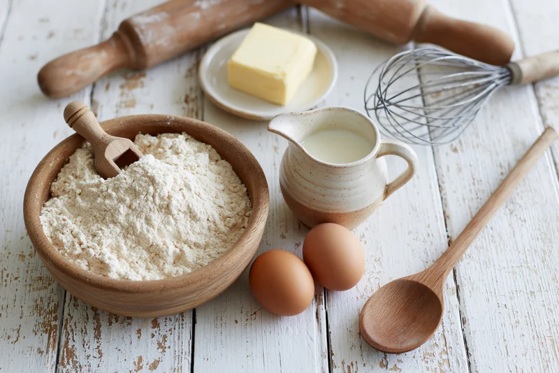 Baking ingredients arranged on a rustic white wooden table with wooden utensils and ceramic pitcher