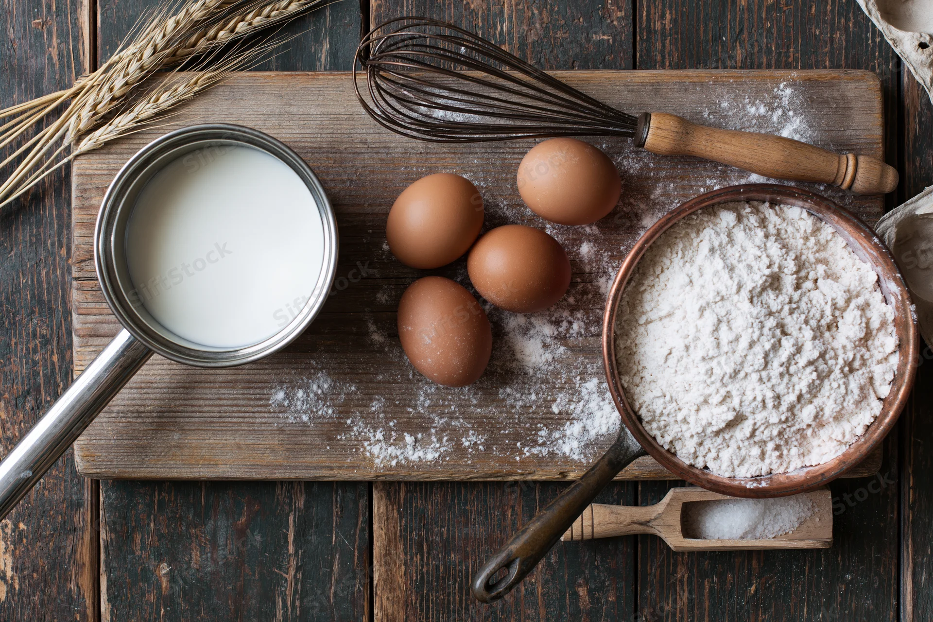 Baking ingredients arranged on a wooden board with flour, eggs, milk, whisk and wheat straw on dark wood surface