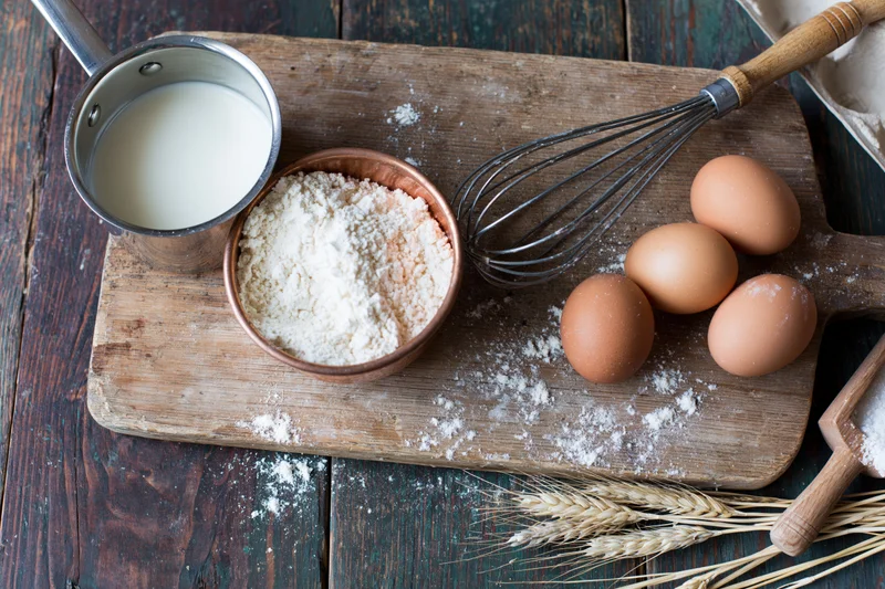 Baking ingredients arranged on a wooden cutting board with flour, eggs, milk, whisk, and wheat stalks