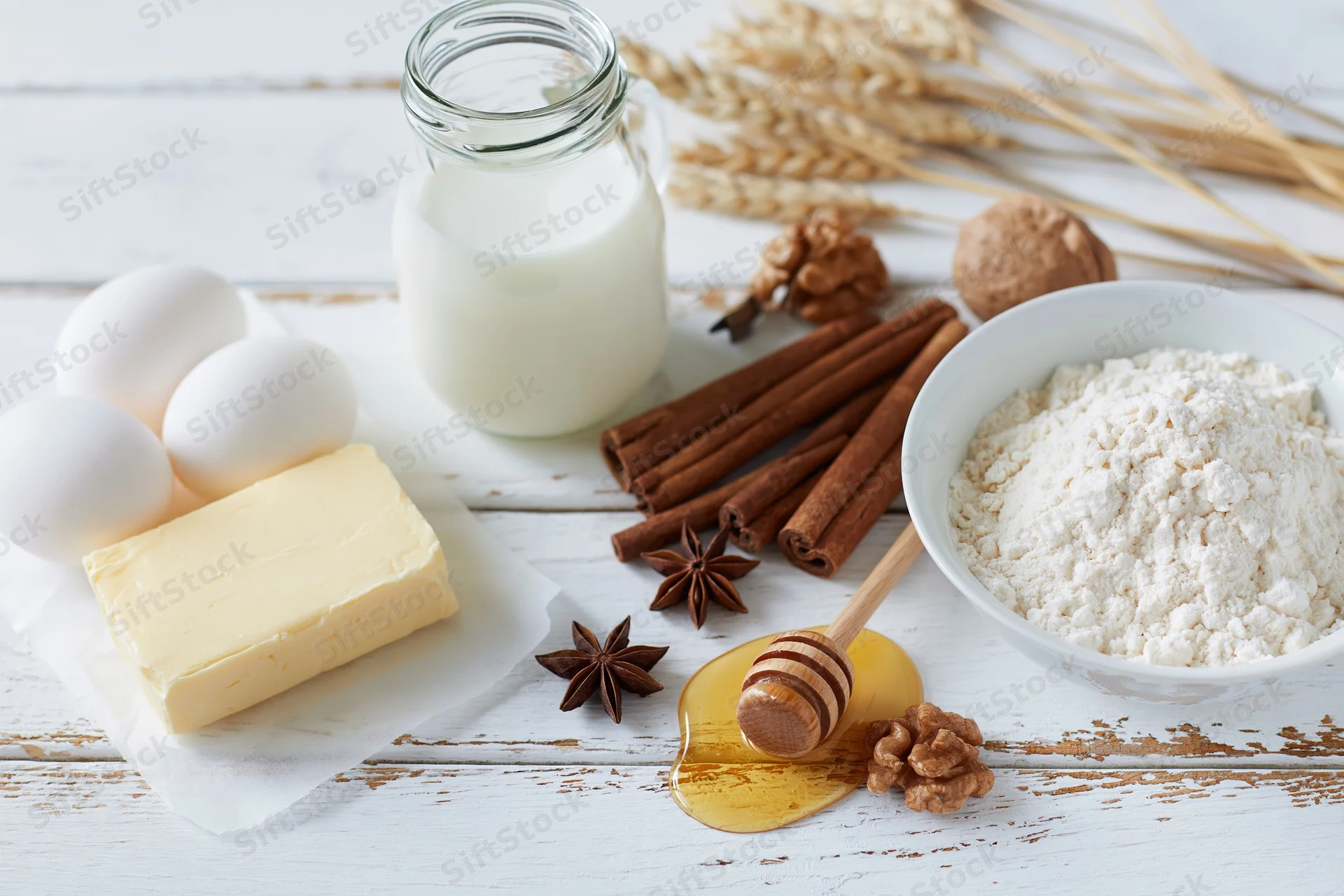 Baking ingredients arranged on a white wooden surface with flour, eggs, butter, milk, honey, cinnamon and walnuts