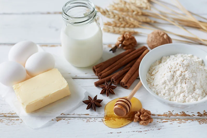 Baking ingredients arranged on a white wooden surface with flour, eggs, butter, milk, honey, cinnamon and walnuts