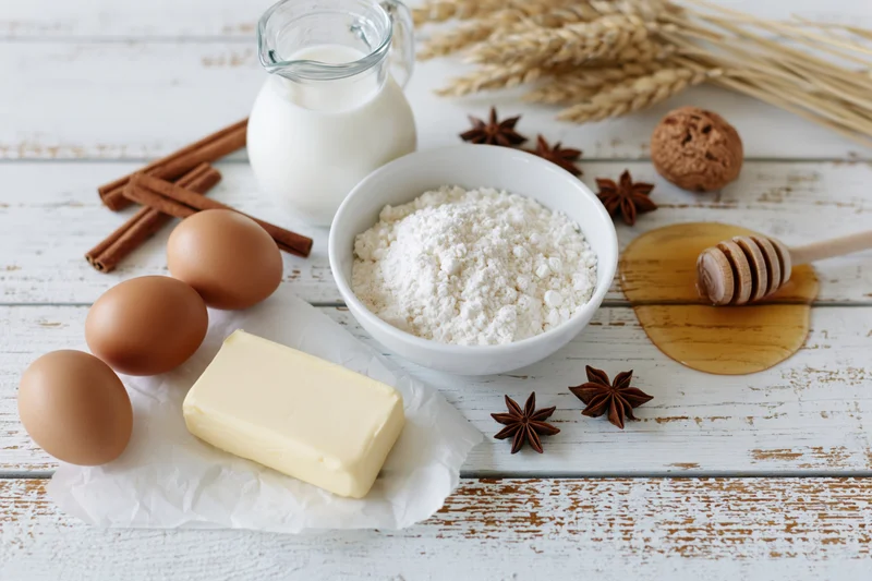 Baking ingredients arranged on a white wooden table with flour, eggs, butter, milk, honey, cinnamon and star anise