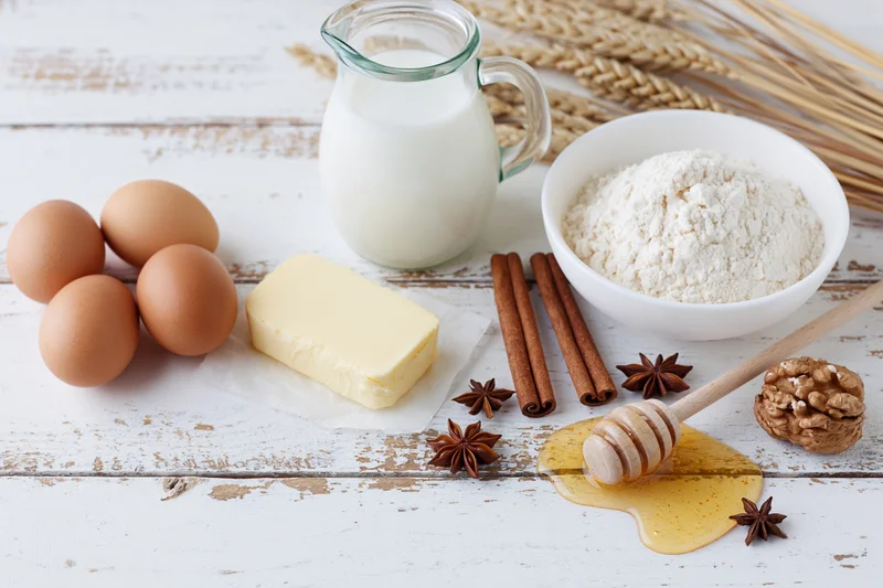 Baking ingredients arranged on a white wooden table with flour, eggs, milk, butter, honey, cinnamon and walnuts