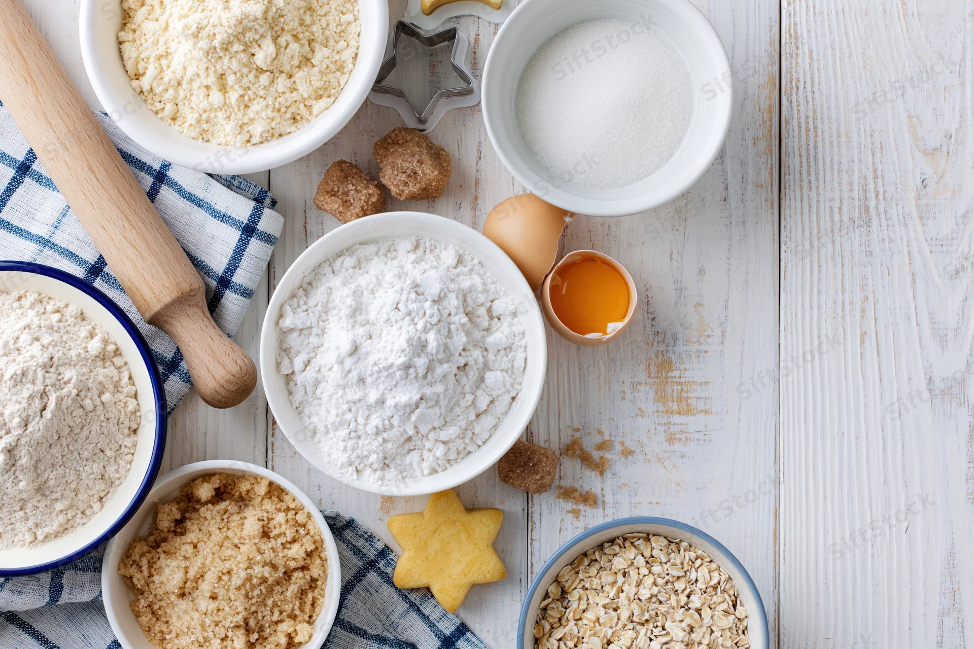 Baking ingredients arranged on a white wooden surface with a star-shaped cookie and rolling pin, including flour, sugar, eggs, oats, and brown sugar in bowls, alongside a checkered cloth and cookie cutter, viewed from above with copyspace on the right