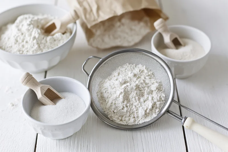 Flour being sifted through a metal sieve on a white wooden table with bowls of sugar and flour nearby