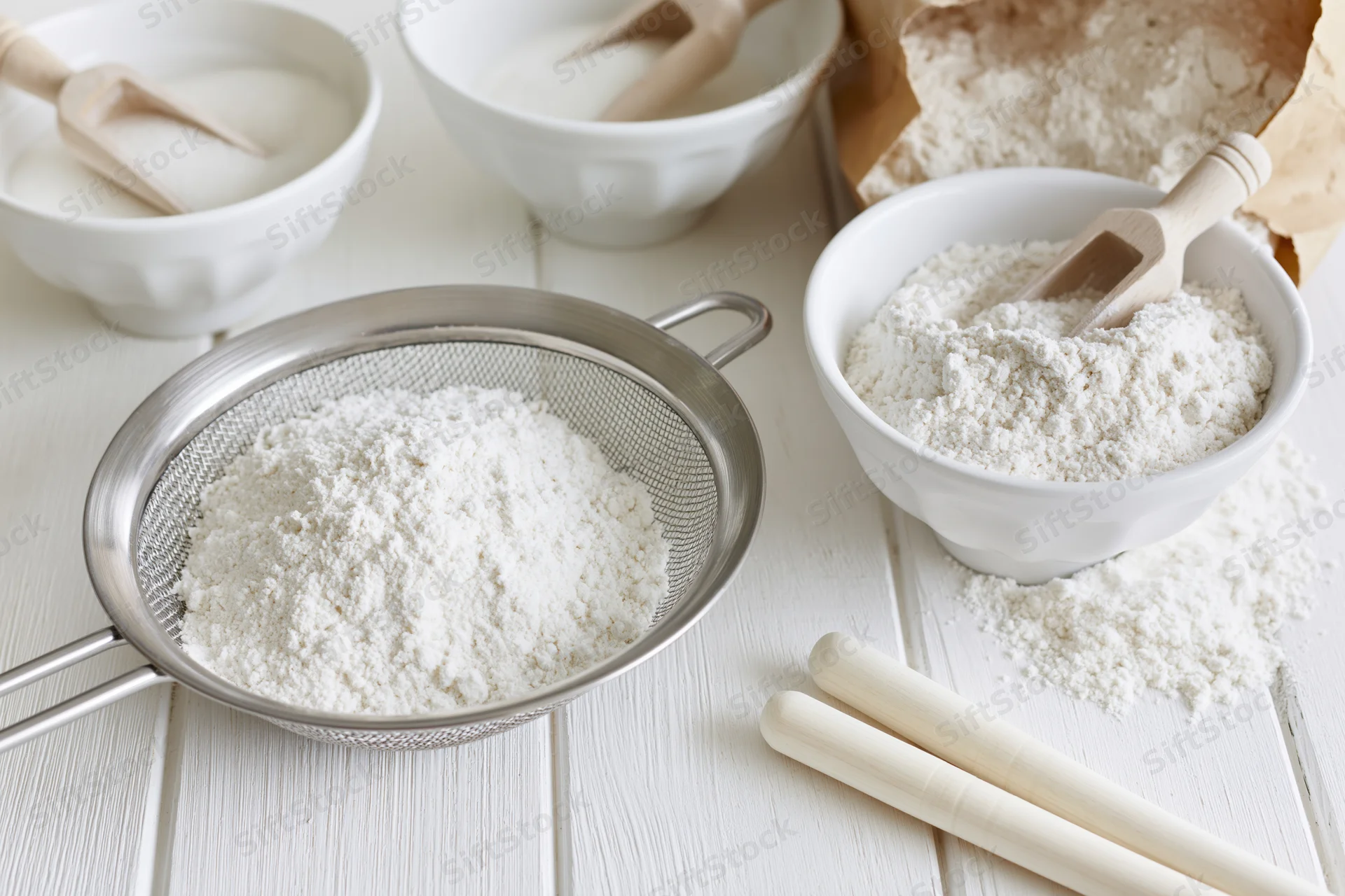 Flour being sifted through a metal sieve on a white wooden table with bowls and wooden spoons nearby