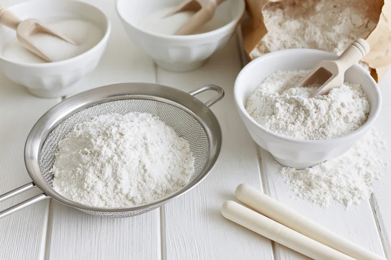 Flour being sifted through a metal sieve on a white wooden table with bowls and wooden spoons nearby