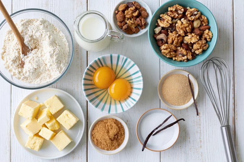 Baking ingredients arranged on a white wooden tabletop with utensils and bowls for recipe preparation