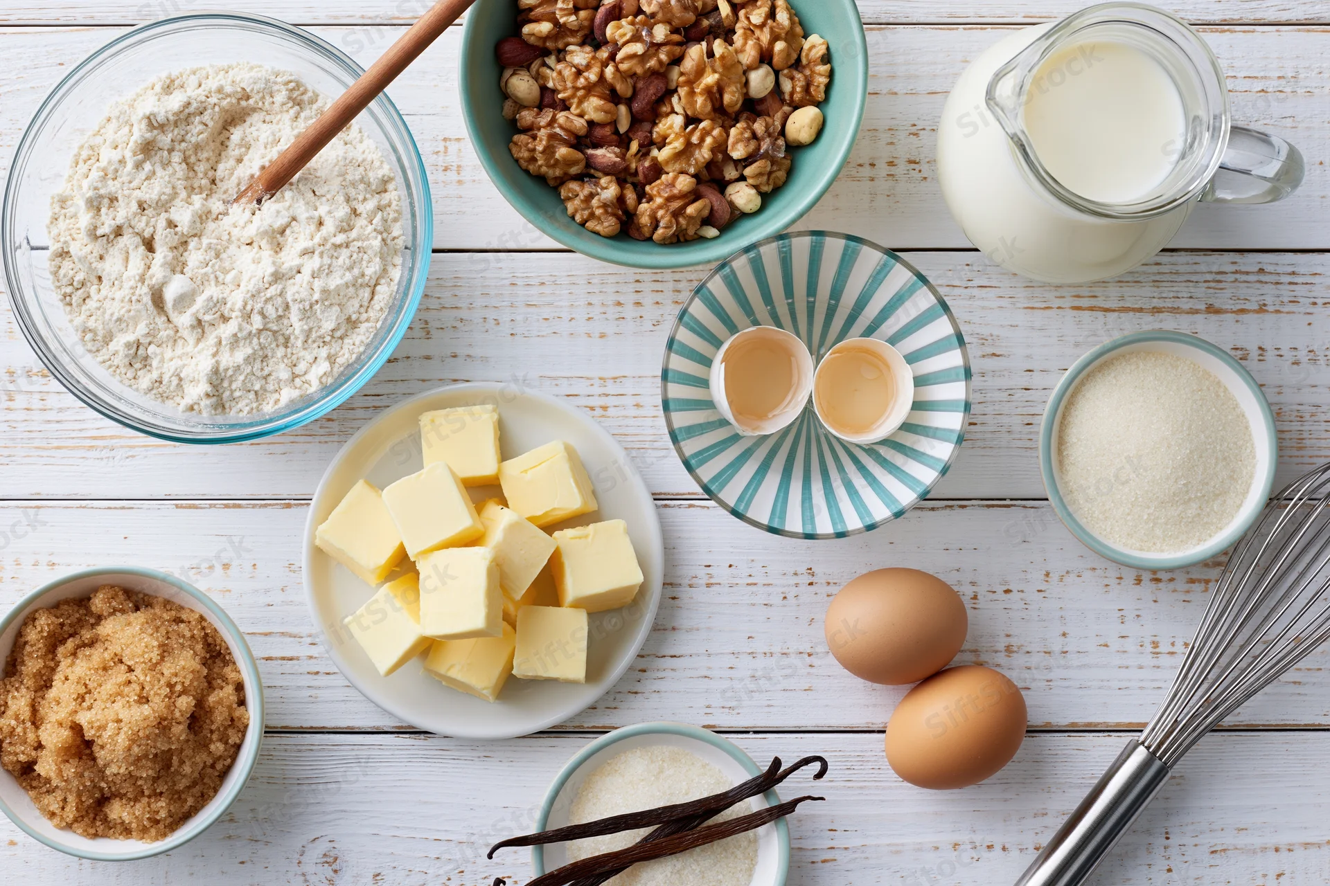 Baking ingredients arranged on a white wooden surface with bowls, eggs, butter, flour, milk, sugar, walnuts and a whisk