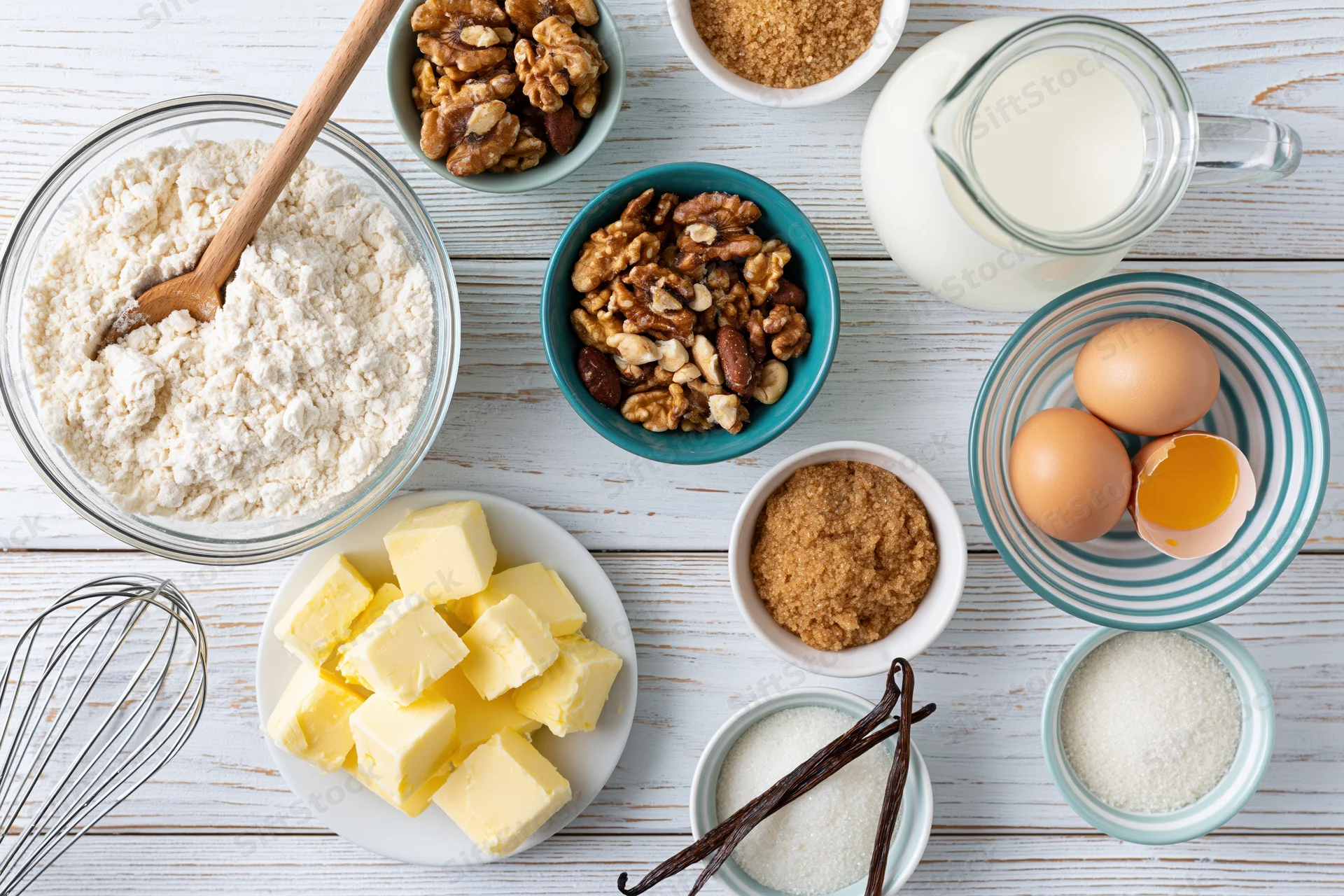 Baking ingredients arranged on a white wooden surface with flour, eggs, butter, sugar, milk, walnuts and vanilla in bowls and containers