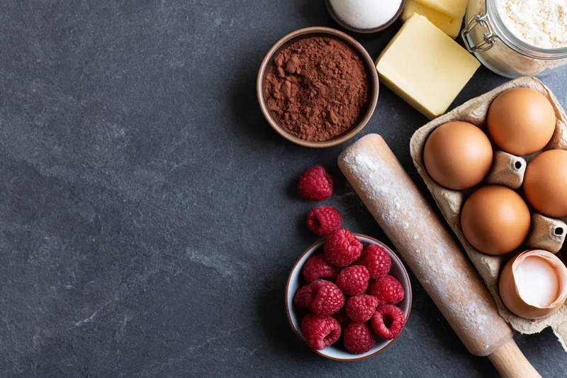 Baking ingredients including chocolate powder, raspberries, eggs, butter and flour arranged on dark countertop with rolling pin and copyspace