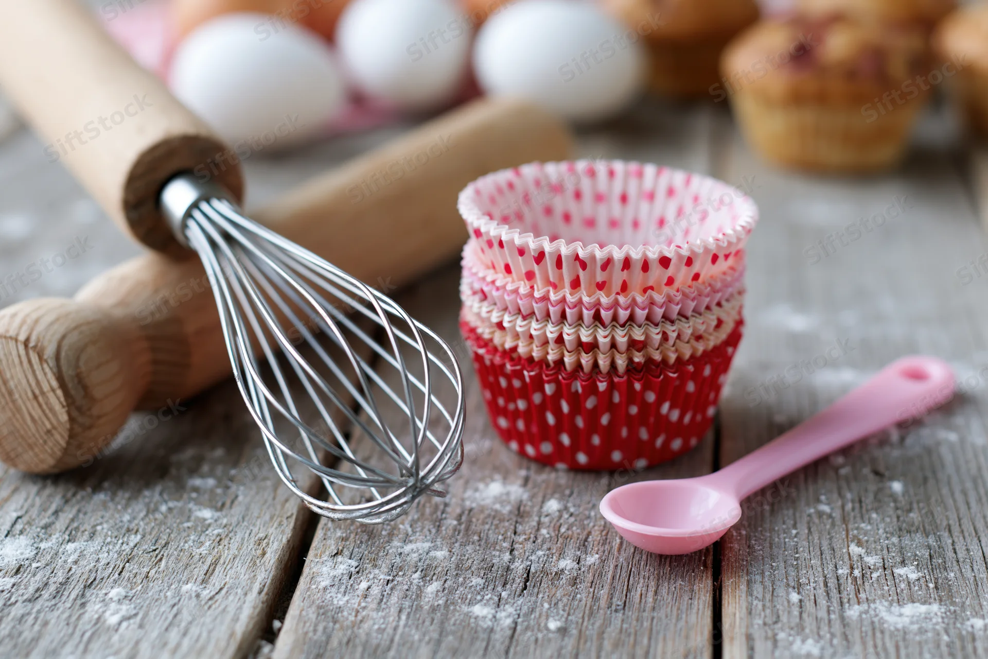 Baking utensils and cupcake liners arranged on a floured wooden table with eggs and muffins in the background