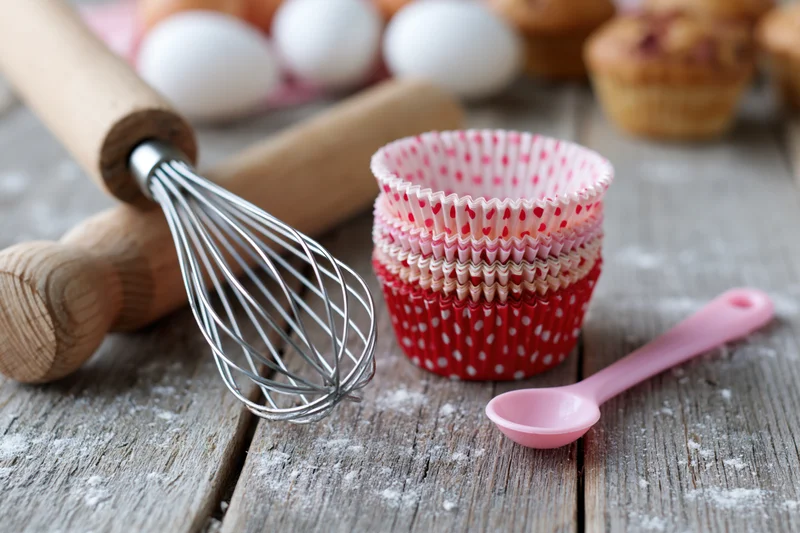 Baking utensils and cupcake liners arranged on a floured wooden table with eggs and muffins in the background