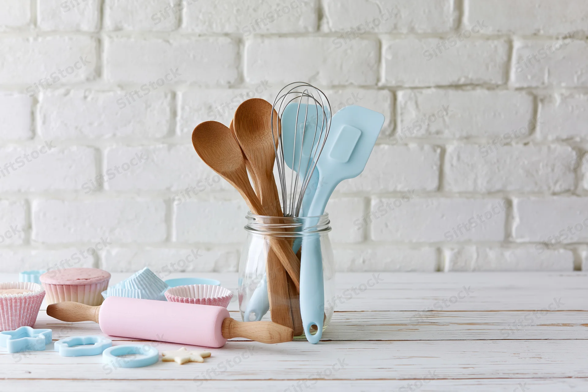 Baking utensils arranged in a glass jar on a white wooden table with pastel-colored tools and cupcake liners nearby