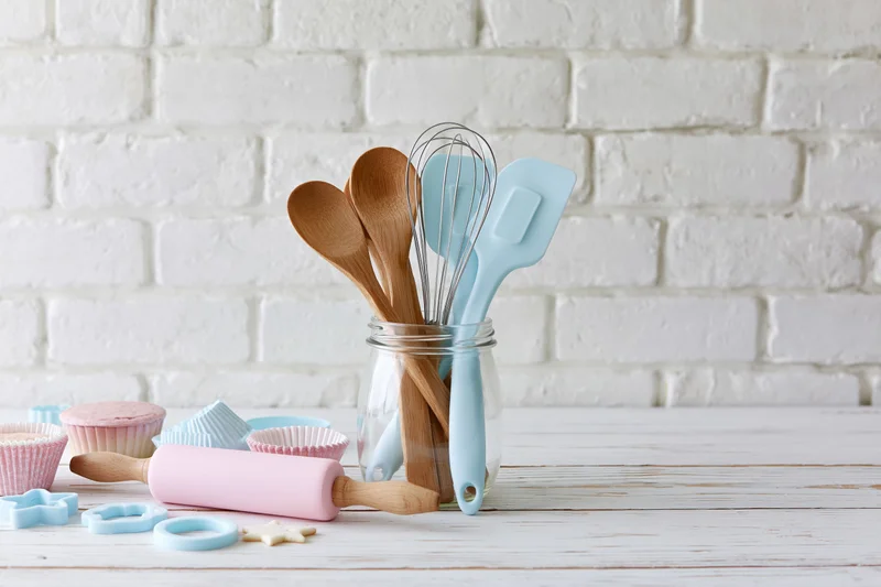 Baking utensils arranged in a glass jar on a white wooden table with pastel-colored tools and cupcake liners nearby
